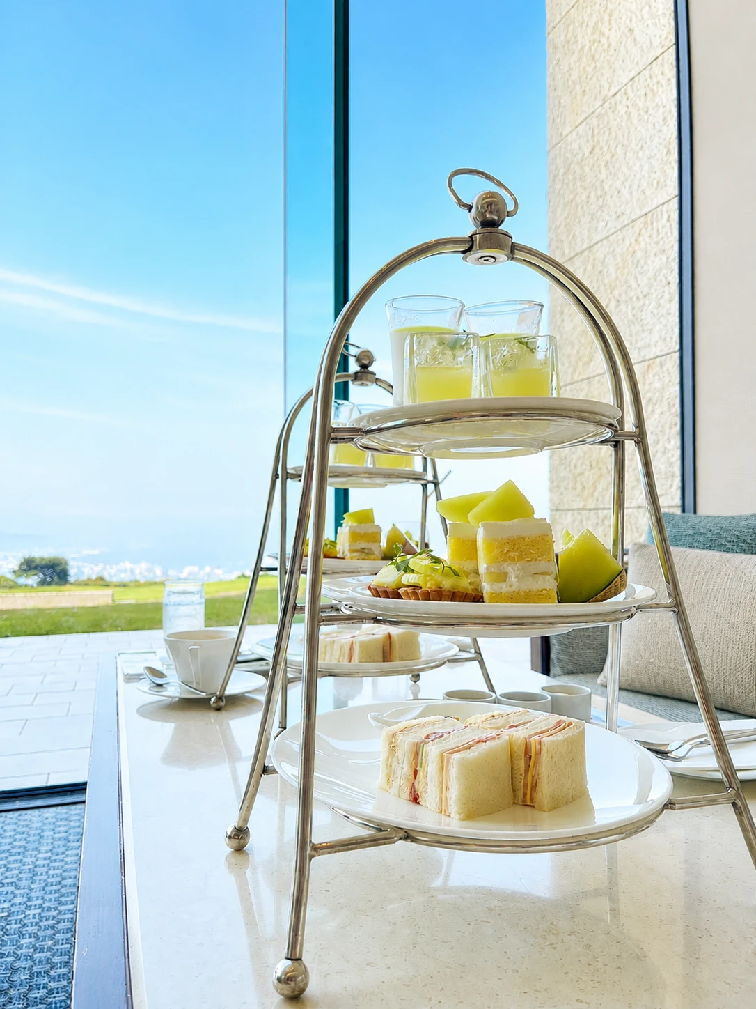 A three-tier silver afternoon tea stand of Crown Melon cakes, tarts and melon wedges, sandwiches and chilled melon juice on a marble table by the lounge window, with a sunlit lawn and the bay beyond